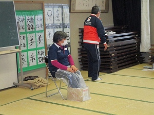 和室で、赤十字のロゴが入ったウェアを着た男女二人がおり、座っている女性は足元をビニールで覆われた箱に足を入れている写真