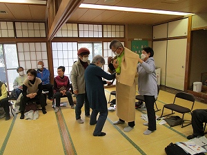 和室で高齢者数名が座って見守る中、男性が着付けの練習台として薄茶色の着物のような衣装を着て、二人の女性から手伝いを受けている様子の写真