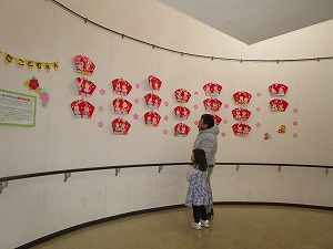 白い曲面の壁にひな人形とピンクの星形飾りが多数貼られた場所で、一人の大人が小さな女の子と一緒に飾り付けをじっと見上げている様子の写