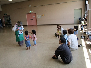 広い部屋の床の上で、エプロン姿の女性が二人の幼児と交流しており、その様子を数人の母親らしき大人が座って見守っている写真