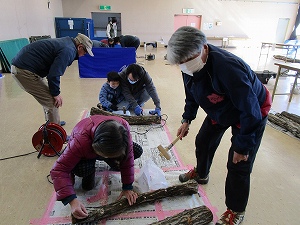 広い室内で、マスクをつけた参加者や子供が、数本の原木に金槌を使ってしいたけの駒打ち作業を行っている写真