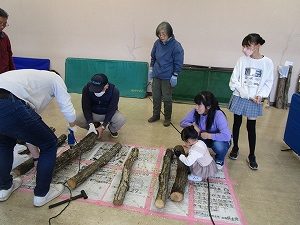 参加者達が新聞紙とピンクのシートが敷かれた床に並べられた数本の原木にしいたけの菌を打ち込んでいる様子の写真