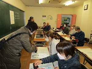 複数人が机に座り机に置かれた新聞紙の上で小さな木に押し込んで入れている様子の写真