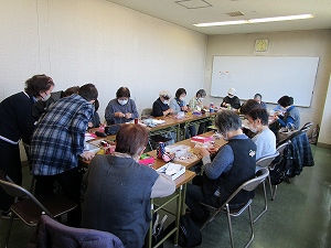 マスクを着用した高齢の女性十数名が長い机を囲み、手芸材料を広げて熱心に手作業に取り組む様子を捉えた集合写真