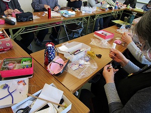 作業中の手元をクローズアップした写真で、布や針、糸、ハサミなどの手芸材料や道具が散乱した机を複数の人が囲み、制作している様子の写真