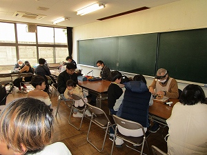 広い教室で、黒板と窓を背にした大人と子供たちが、マスク姿で机を囲み、老眼鏡のようなものをかけた男性がおもちゃを修理している写真