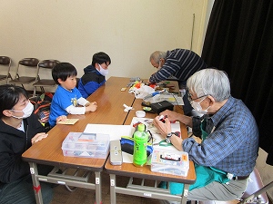 長机を囲んで大人と子供が向かい合い、手前では老眼鏡とマスクをつけた高齢の男性がおもちゃを修理しているのを子供が見ている写真