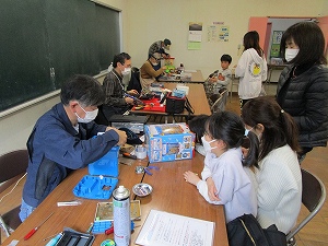 大人と子供が机を囲み、男性が青い筐体の電子機器のようなものを分解している様子を二人の子供と大人が興味深そうに見つめている写真