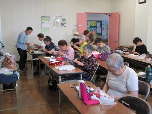 十数名の女性たちが机に向かい、講師の指導のもとねこのストラップ作りに熱心に取り組んでいる様子の写真