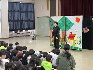 広間で、大勢の子供たちが座って見守る中、手作りの舞台装飾を背景に黒い服を着た女性が話をしている様子の写真