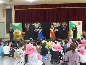 広間において、多数の子供たちが飾り付けがされた舞台を囲み、布製の人形を使った人形劇を鑑賞している様子の写真