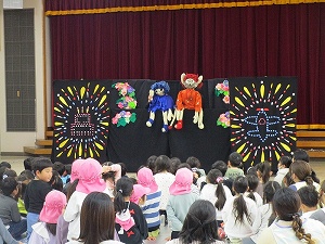 舞台の下で、黒のパネルに花火や飛行機のような絵が描かれ二体の大きな人形が座っているのを、床に座った大勢の園児や子供たちが鑑賞している様子の写真