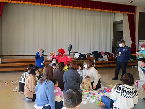 ステージの前で、鬼の面をつけた赤い服と青い服を着た人が中央に座り、たくさんの親子連れが座ってボールやおもちゃで遊んでいる写真