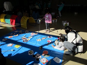 光の差し込む広い室内で、青いシートの上でキャラクターの絵柄の魚などを釣り上げる遊びを、子供と大人が一緒に楽しんでいるイベント風景の写真