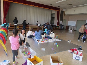 広い会場で、複数の親子がレジャーシートの上でボール遊びやトンネル遊びをしたり、ステージの周りでくつろいだりしている自由遊びの様子の写真