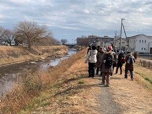 冬枯れの河川敷の土の道を、ダウンジャケットや厚手のコートを着た数人の人々が、川の流れと住宅街を右手に眺めながら奥に向かって歩いている様子の写真
