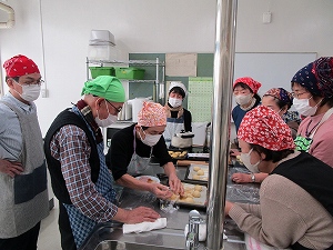 白とステンレスが基調のキッチンで、バンダナやエプロン、マスクを着用した男女が集まり、女性がステンレスの作業台の上で生地を丸めるパン作りの講習風景の写真