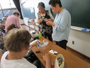 教室内で数名の女性が長机を囲み、手前の女性が手芸道具を準備し、奥の二人が立ってお互いの手の動きを見ながら熱心に手芸の細かい作業について意見交換をしている様子の写真