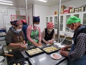 厨房内でバンダナ、エプロン、マスクを着用した高齢者が集まり、参加者が生地をのばし、女性が丸めたパン生地に卵黄を塗るなど調理実習をしている様子の写真