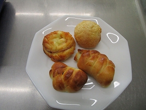白色の八角形の皿に、丸いメロンパンのようなものと惣菜パンのようなものが計四つ盛り付けられている、調理実習の成果であるパンのアップ写真
