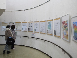 緩やかなカーブを描く白い壁に、ワイヤーで吊るされた複数のポスターやイラスト作品が展示されており、2人の女性がそれらを鑑賞している様子の写真