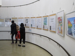 螺旋状の手すりが続く白い壁面で、文字情報やイラストが描かれた複数の額装された作品がワイヤーで吊るされて展示されており、2人が鑑賞している様子の写真