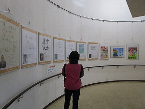 白い曲線状の壁にワイヤーで吊るされた複数のポスターやイラスト作品を、女性が熱心に鑑賞している様子の写真