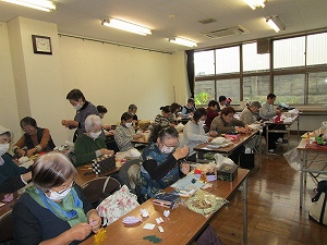 大きな窓がある明るい教室で、マスクを着用した高齢者が机に向かい、それぞれ手芸材料を広げて熱心に作業に取り組む集まりの様子の写真