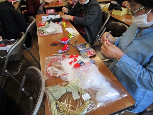 作業中の手元を写した写真で、マスクを着用した女性が赤い布や白い綿、しめ縄状の飾りなどの手芸材料や道具を机に広げ、熱心に作業をしている様子の写真