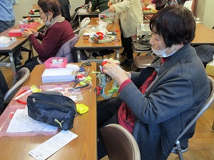 広い会場でマスクを着用した女性たちが机に向かい、手前の一人が赤い飾りを縫い付けている真剣な表情の手芸作業風景の写真