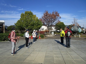 青空の下、広場のタイル敷きの地面で、ポールウォーキング用の杖を持った高齢者達が指導を受けている様子の写真