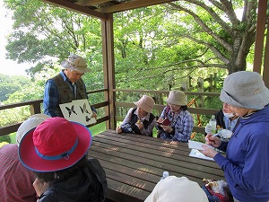 山の緑を背景にした屋外の休憩所で、数名の参加者がテーブルを囲み、帽子を被った指導者らしき人物がチョウのシルエットが描かれた紙を使いながら自然観察のレクチャーを行っている屋外写真