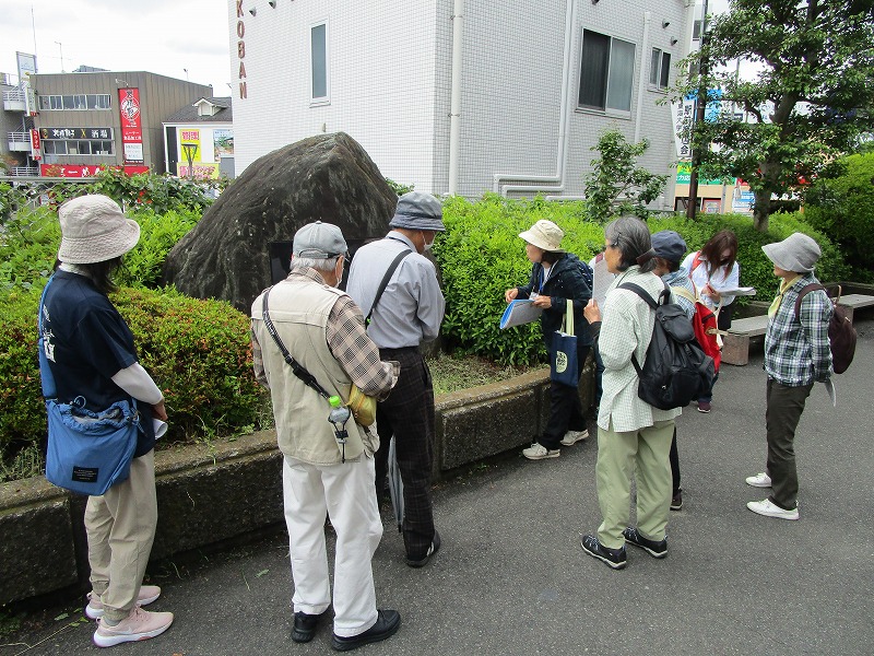 都心のアスファルト舗装された広場で、帽子を被った数名の参加者が、大きな自然石と植え込みのそばで、説明を聞いている写真