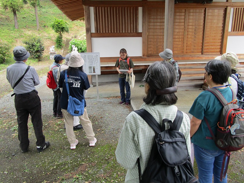 木造の建物の軒下で資料を手に説明する女性ガイドの話を、リュックを背負い帽子をかぶったハイキングスタイルの参加者たちが取り囲むようにして聞いている写真