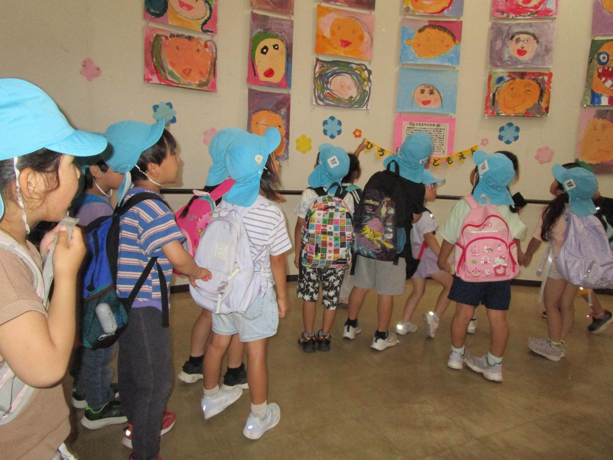 水色の帽子とリュックサックを背負った複数の子供たちが、壁一面に展示された子供たちの描いた鮮やかな人物画の絵の前を、列になって歩きながら鑑賞している様子の写真