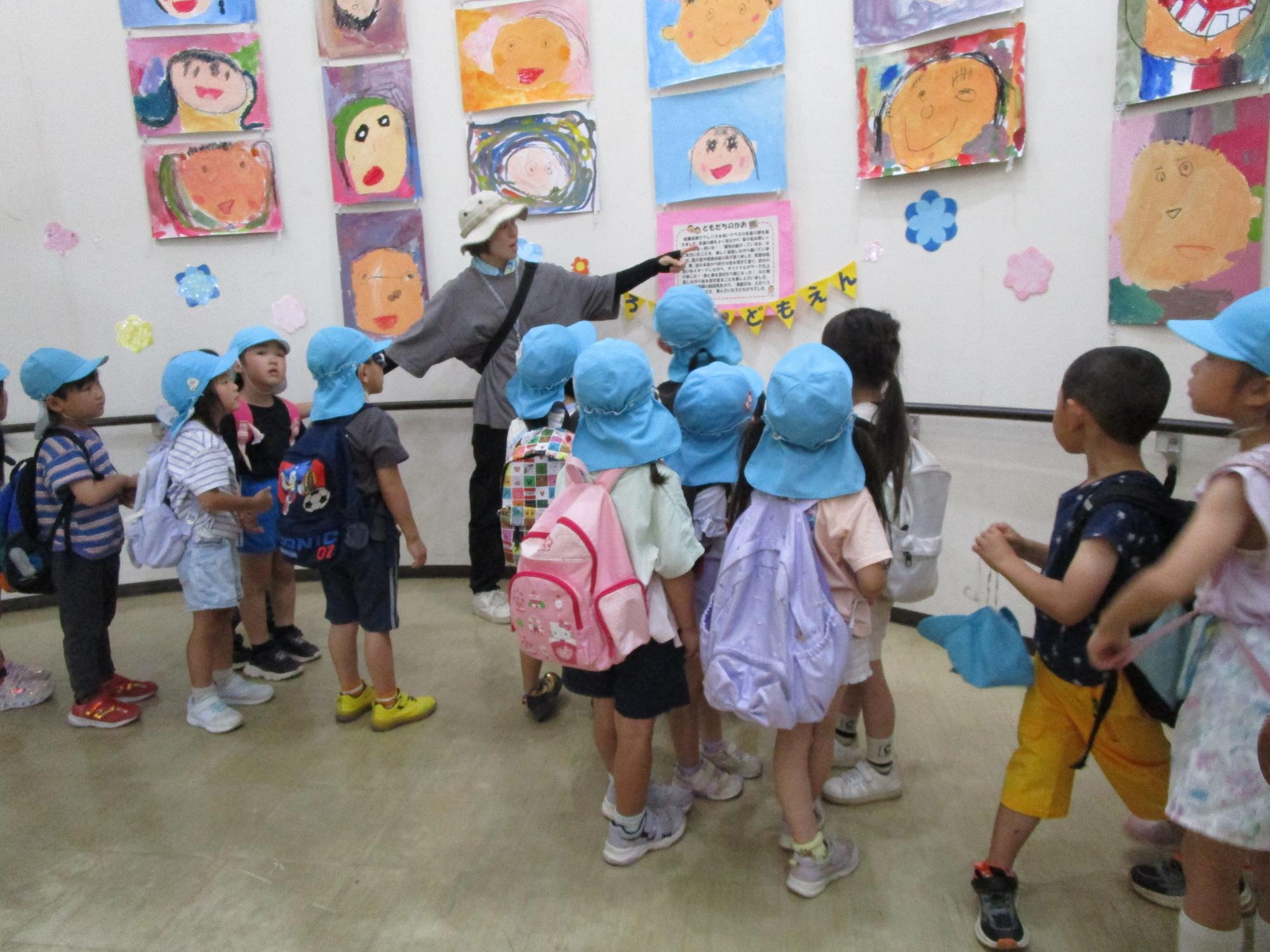 水色の帽子とリュックサックを背負った幼児たちが、壁一面に展示された子供たちの描いた鮮やかな人物画の絵の前で、引率の先生の説明を聞きながら鑑賞している様子の写真