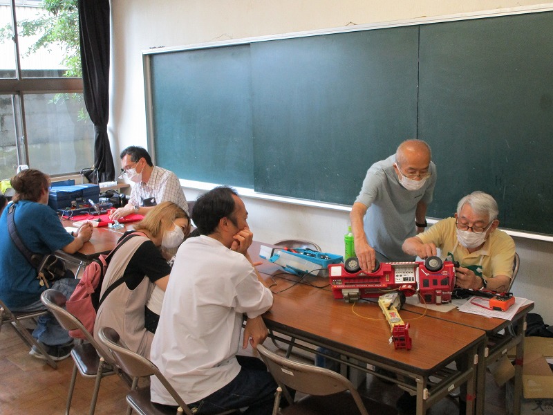 地域住民が持ち寄った壊れたおもちゃを、高齢のボランティアが机の上で工具を使って修理している様子を、他の住民が見守っている風景の写真