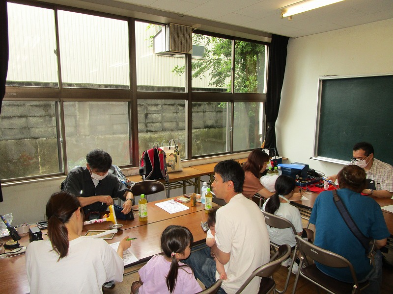 明るい教室で、地域住民が子ども連れで参加し、机を囲んでおもちゃなどの修理を見学・相談している様子の写真
