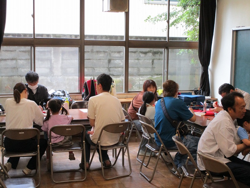 地域の親子連れが明るい教室で修理ボランティアと一緒に壊れたおもちゃを囲みながら相談したり見学したりしている様子の写真