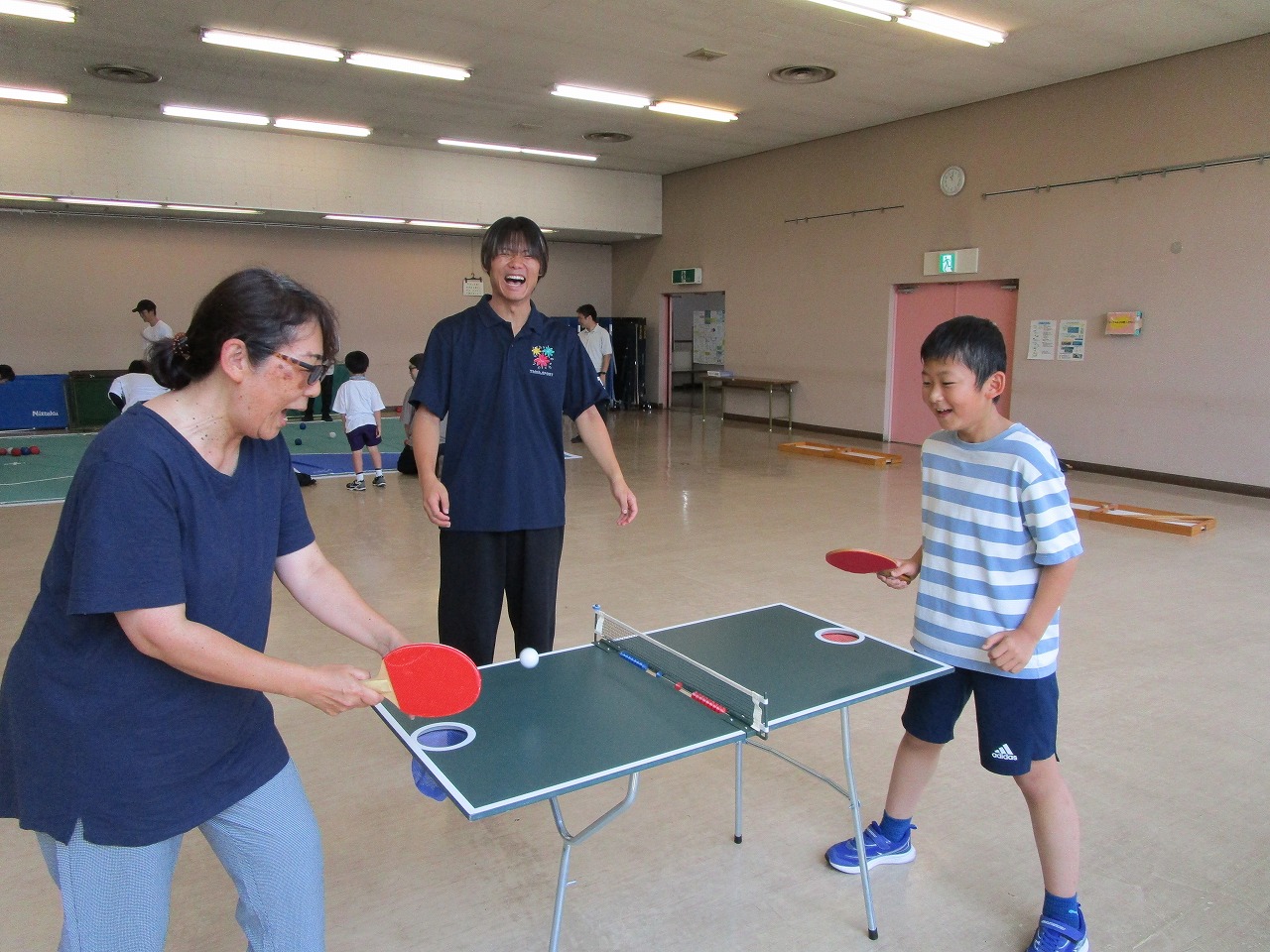 体育館のような広い屋内施設で、卓球台のような小さな台を使って、笑顔の女性と少年がラケットとピンポン玉で卓球を楽しんでいる写真