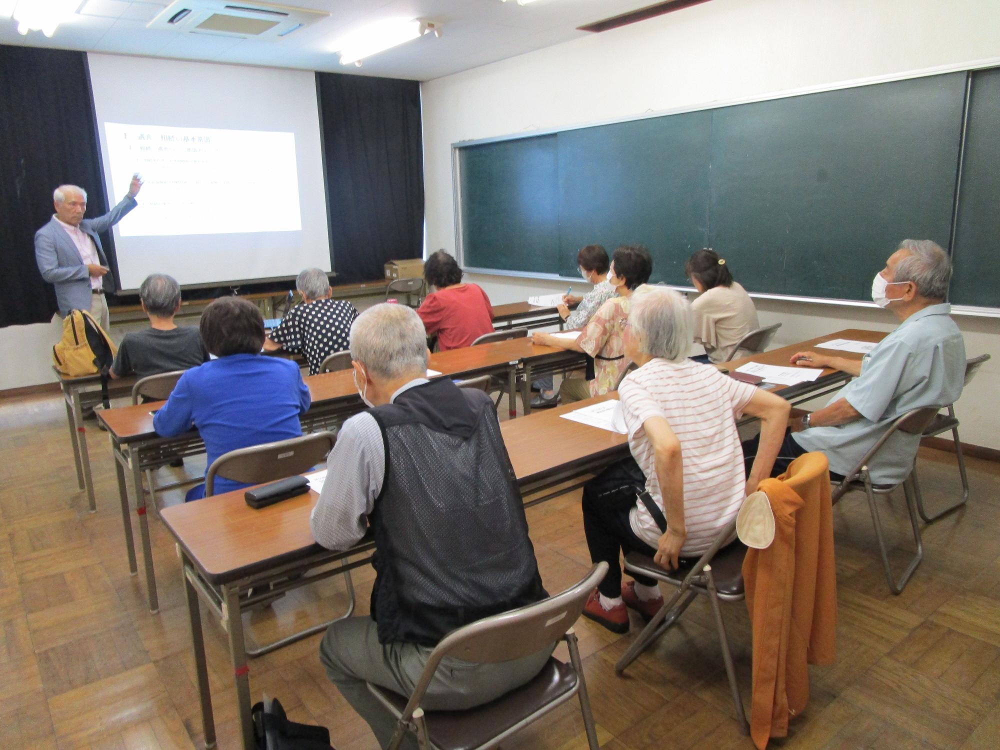 木の床と黒板がある教室で、中高年の参加者十数名が机に向かい、講師の男性がプロジェクターのスクリーンを指差しながら講義を行っている写真