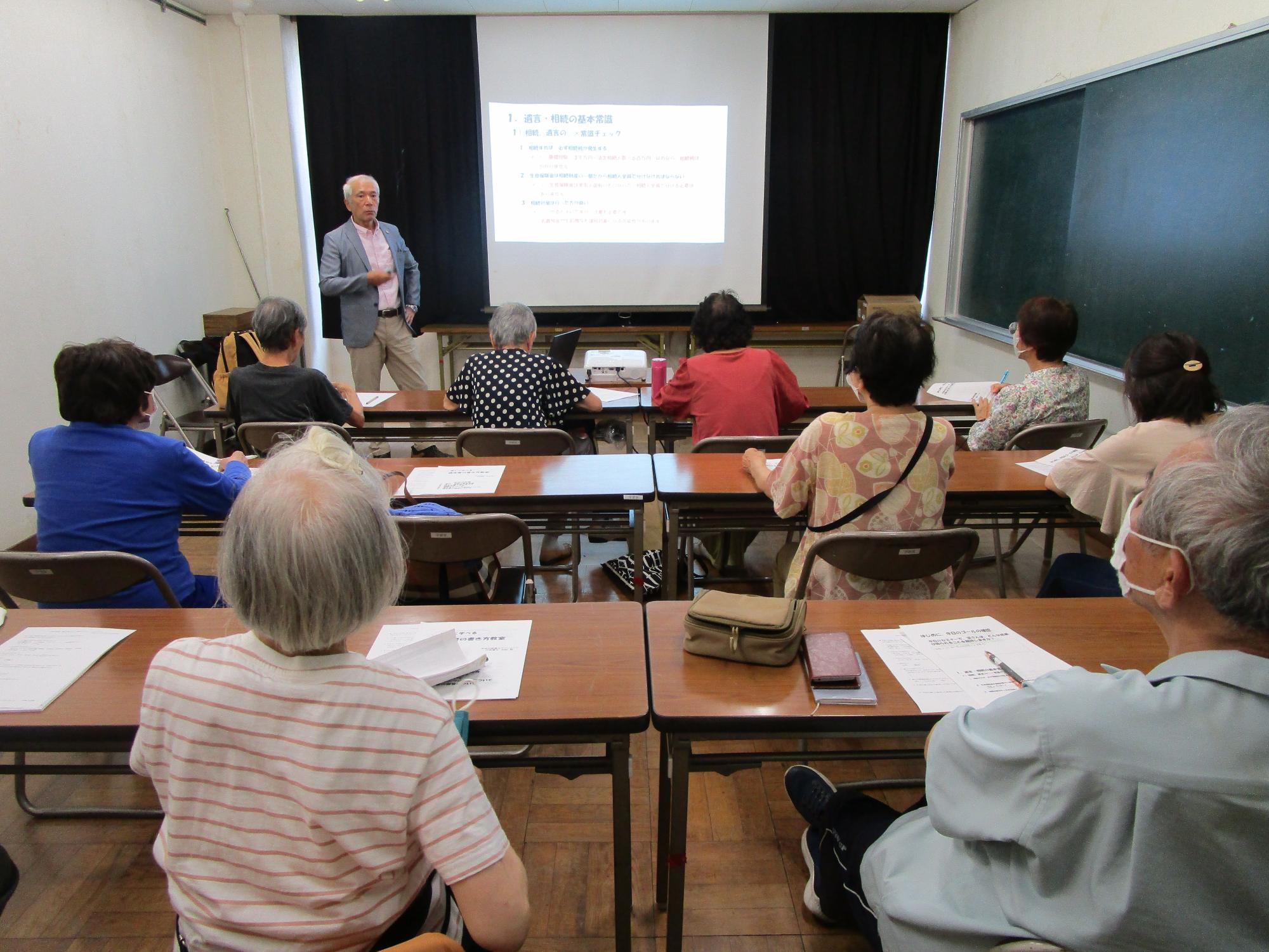 教室の後方から撮られた写真で、数名の中高年の参加者が机に座り、前方の講師の男性がプロジェクターに映し出されたスライドを使いながら進めている講習会に参加している様子の写真