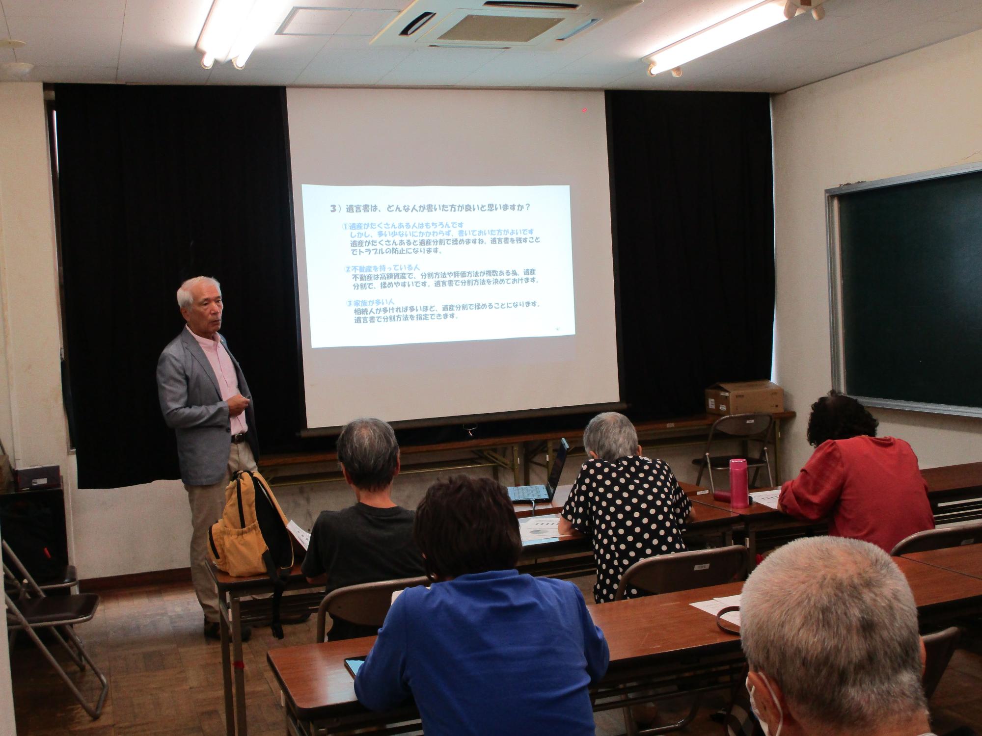 男性講師がプロジェクターのスクリーンに映し出されたテキストを指し示しながら、中高年の受講者十数名に向けて講義を行っている遺言書の書き方セミナーの写真