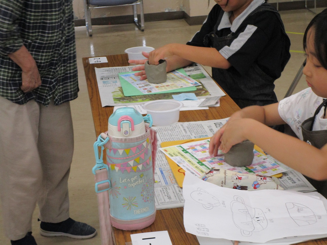 机を囲むように座った子供たちが、新聞紙の上に置かれた紙粘土のような素材を熱心にこねて形を作り、傍らに立つ大人がそれを見守っている様子のクローズアップ写真