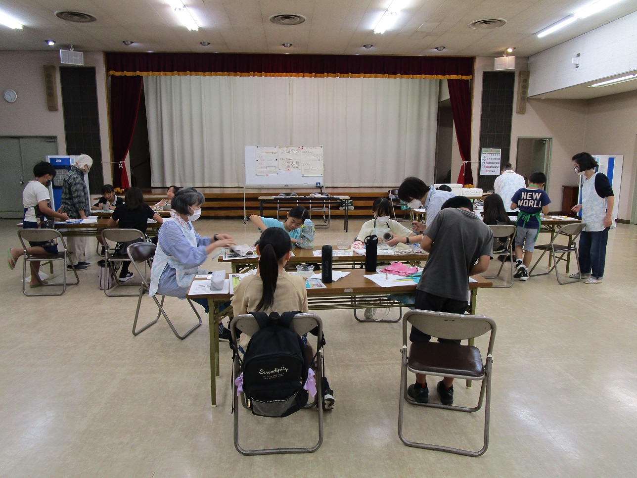 数人の子供たちと大人が長机に分散して向かい合い、それぞれが作業に取り組んでいる写真