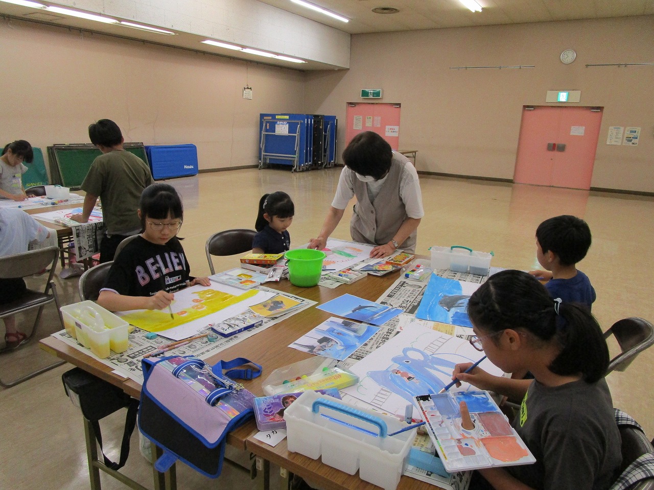 地域の広い集会室のような場所で、数人の子供たちがテーブルを囲み、エプロン姿の女性に教わりながら、新聞紙を敷いた上で絵の具を使って熱心に絵を描いている様子の写真