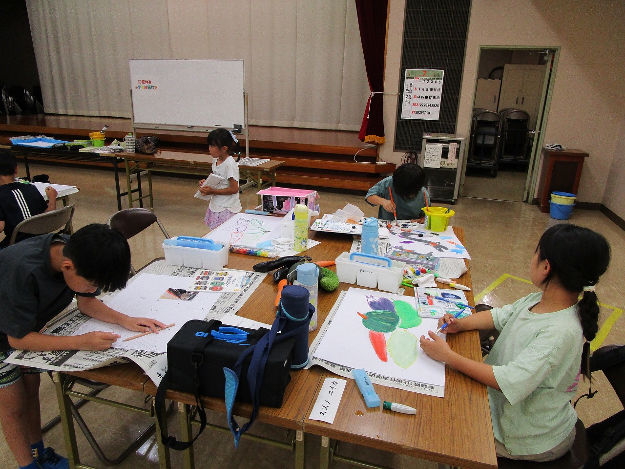 数人の子供たちがテーブルを囲み、新聞紙を敷いた上で絵の具や色鉛筆を使い、絵を熱心に描く絵画教室の写真