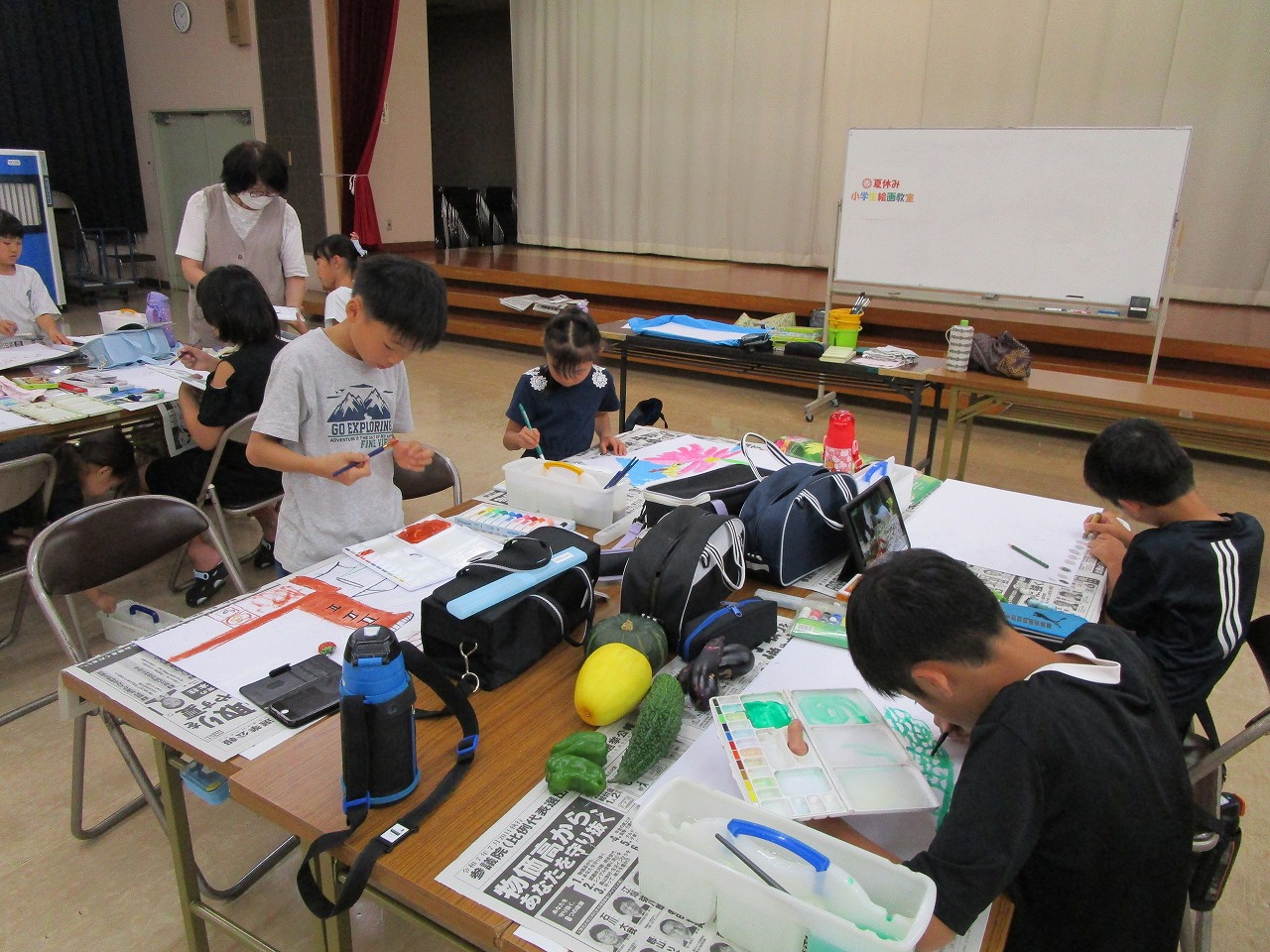 エプロンとマスク姿の女性講師が見守る中、子供たちが新聞紙を敷いた机の上で絵を描いている絵画教室の写真