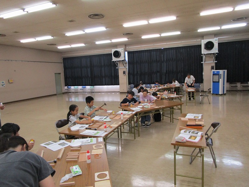 多くの子供たちが木工や工作の体験教室に参加し、長机に座って金槌や接着剤を使いながら熱心に作業に取り組んでいる様子の写真