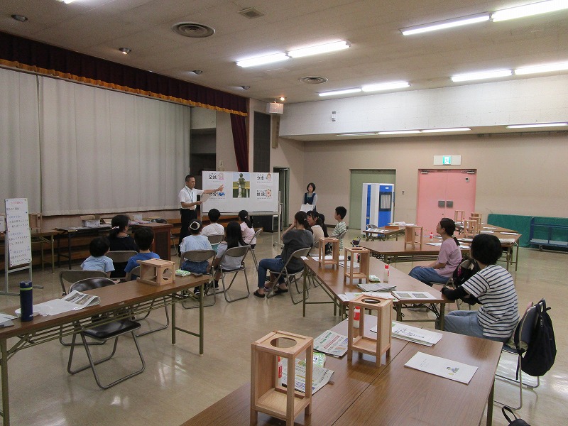 男性が黒板の代わりに置かれたパネルを指差しながら、木工教室に参加した子供たちと数名の保護者に向けて作業の説明をしている様子の写真